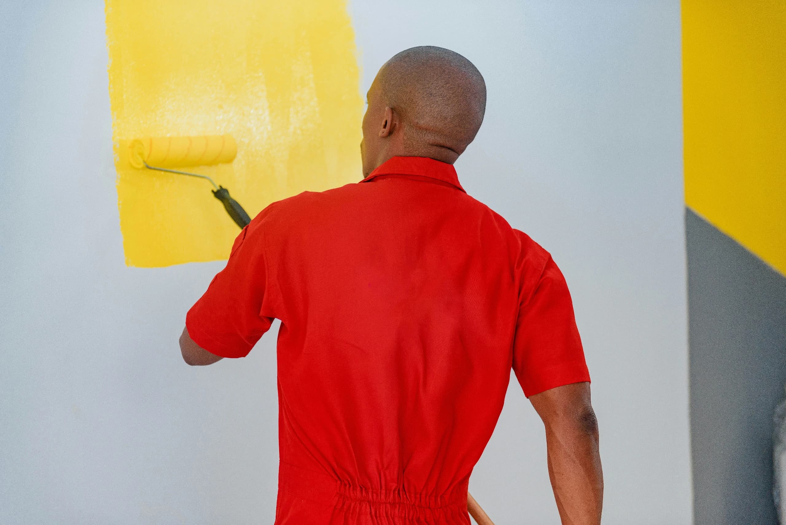 Black man, wearing bright red shirt, painting a white wall yellow
